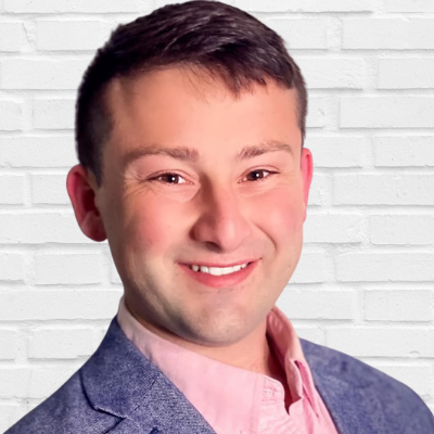 A photo of Aaron Welcher the Communications Director smiling while wearing a blue suit with a pink button up shirt. He is standing in front of a white brick wall.